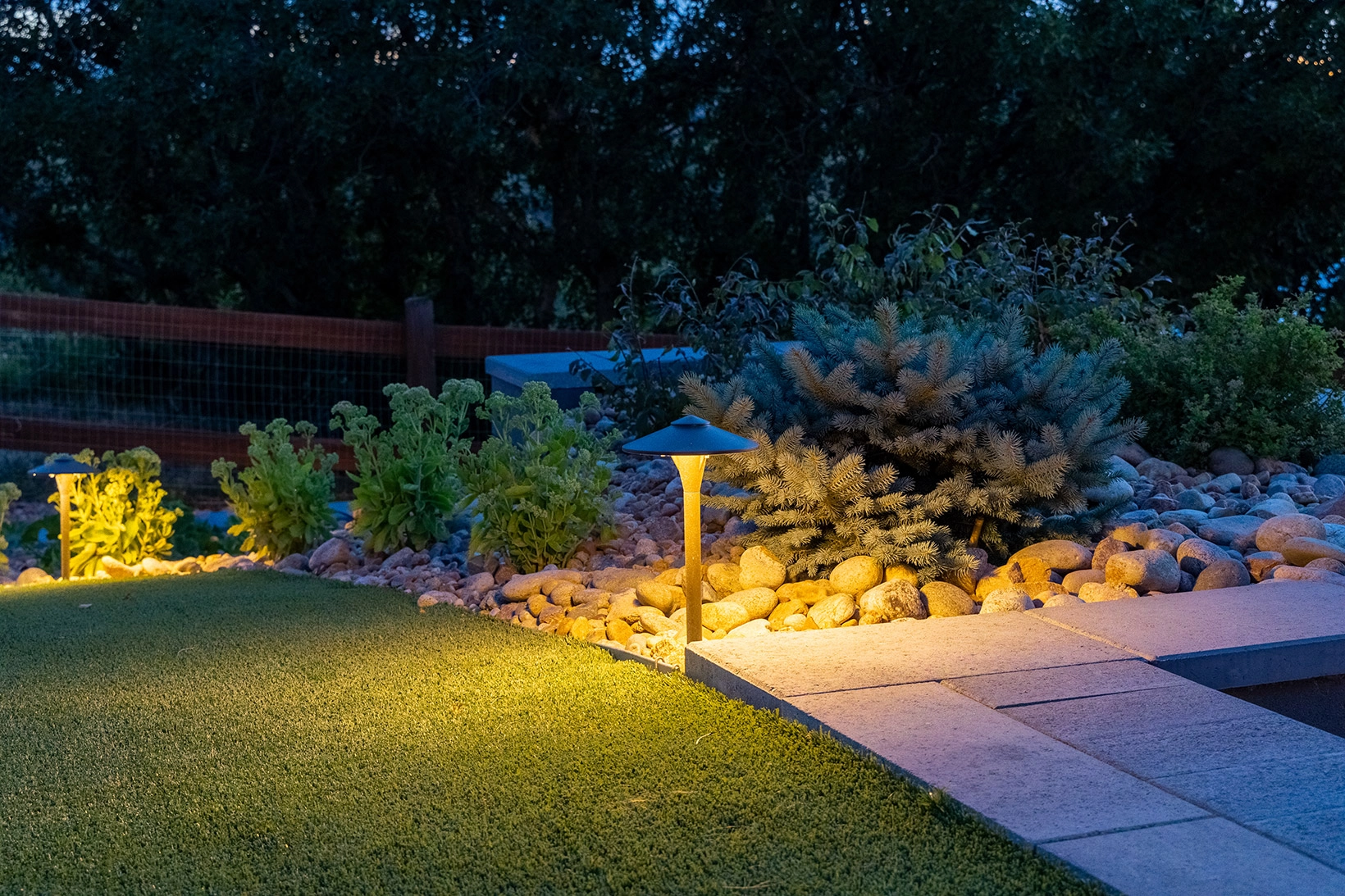 Landscape path lights illuminating a stone walkway and river rock garden bed at dusk