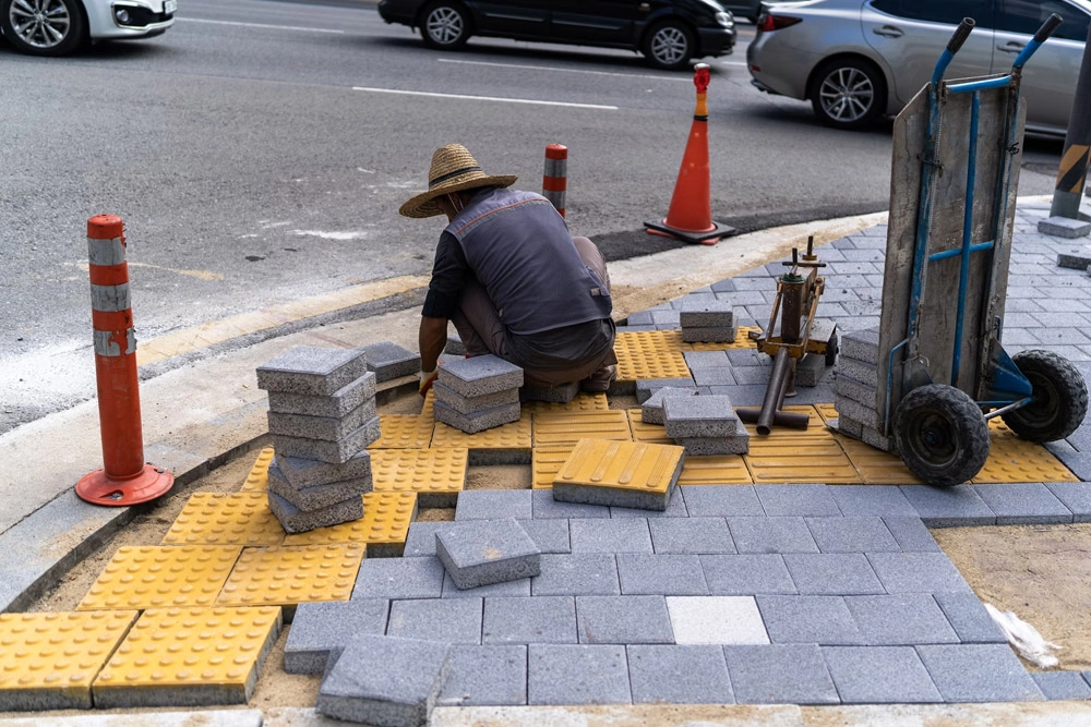 Worker installing gray pavers and tactile paving on a sidewalk corner