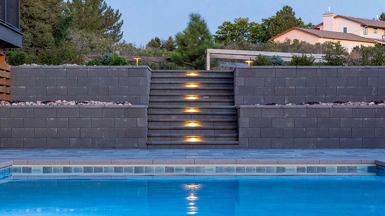 Illuminated staircase between block retaining walls beside a pool at dusk